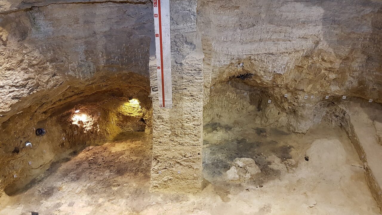 Coupe de fouille dans un abri sous roche, avec une échelle graduée de lettres posée contre une colonne de sédiments et des repères numérotés au sol.