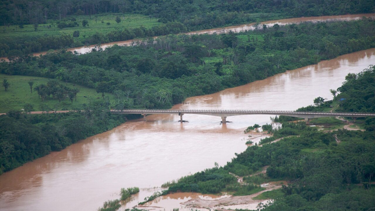 Vue aérienne d’une rivière boueuse traversée par un pont, entre des rives couvertes de forêt tropicale.