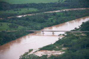 Vue aérienne d’une rivière boueuse traversée par un pont, entre des rives couvertes de forêt tropicale.