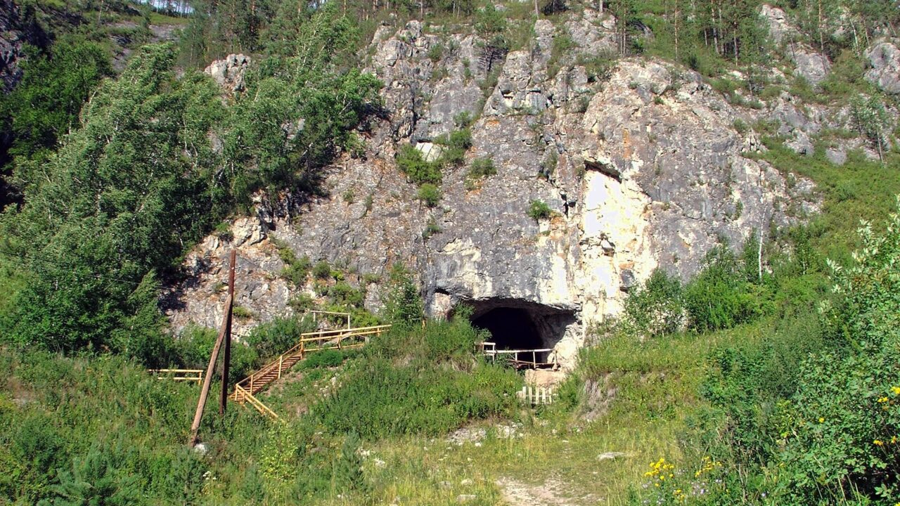 Entrée sombre d’une grotte au pied d’une falaise calcaire boisée, accessible par un sentier et un escalier en bois.